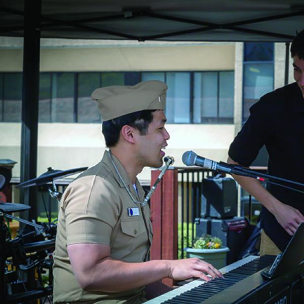 A man in brown Navy uniform plays the piano and wears a harmonica on a neckstrap while another man looks on