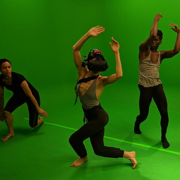 A dancer wearing virtual reality headgear performs with other dancers in a green room