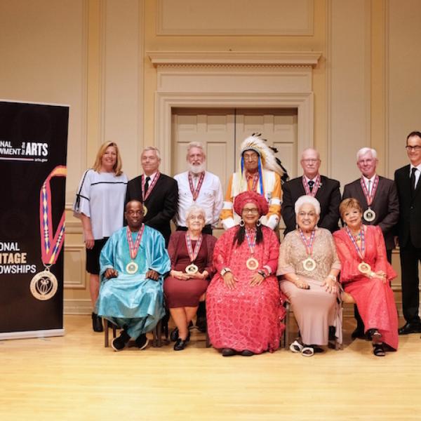 Group of 12 men and women pose for the camera on a stage with two banners saying "National Endowment for the Arts." The front row is seated and the back row is standing.