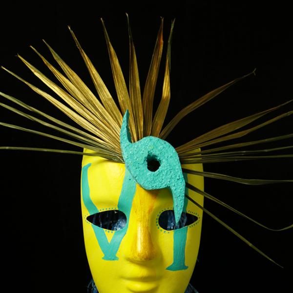 A yellow mask with "VI" painted on it in teal with gold palm fronds branching off the top and a teal hurricane eye on one side