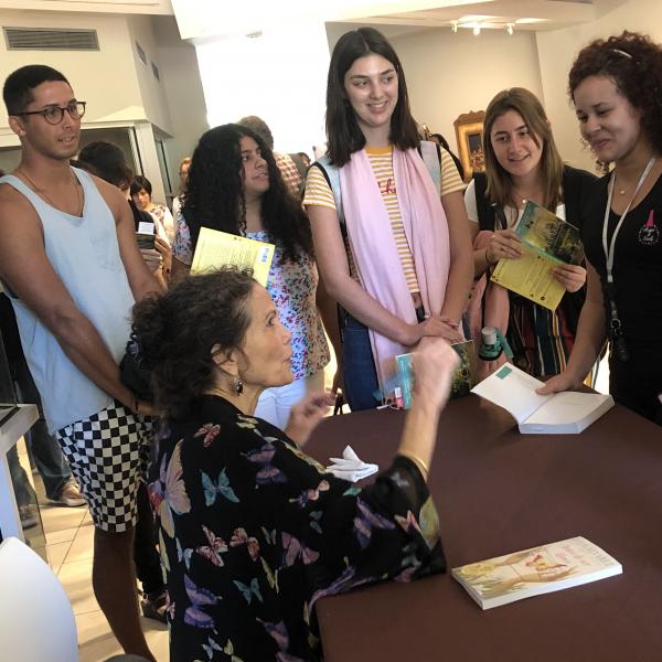 A woman sits behind a desk signing books with a crowd of people around her. 