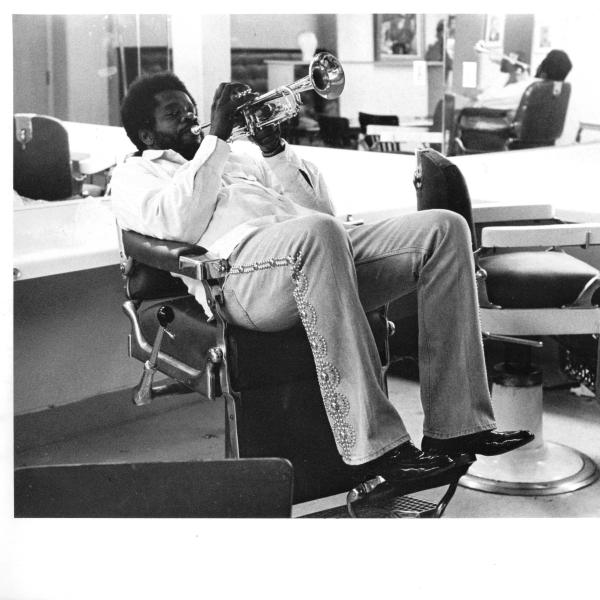 Man sitting in barber chair blowing a trumpet. 