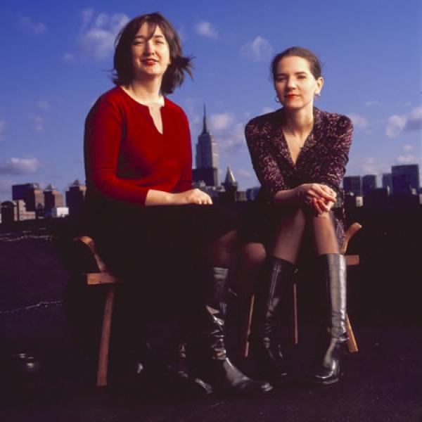 Two women seated on a rooftop open sky and clouds and the city skyline in the background.