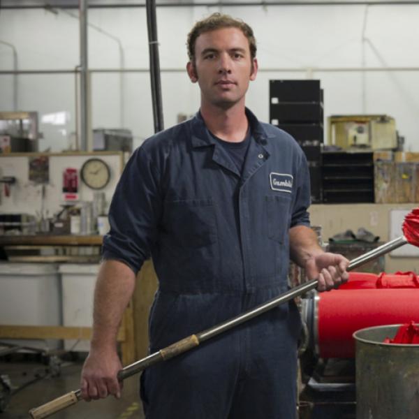 Man standing in front of paint rolling machinery holding a pole with red paint on end. 