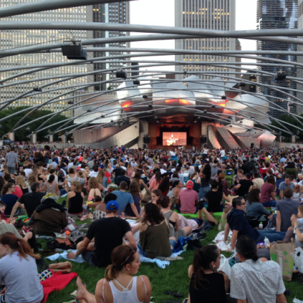 The silver Pritzker Pavilion in with audience in front sitting on grass