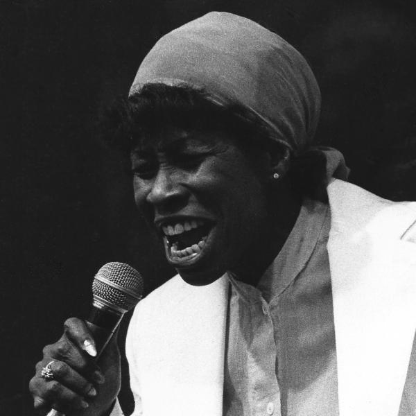 Woman with kerchief on her head singing into microphone. 