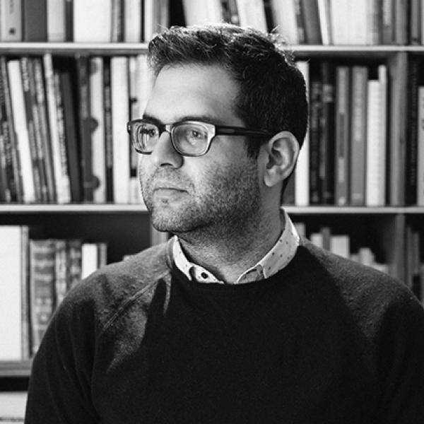Portrait of man wearing dark glass frames in front of a bookcase. 