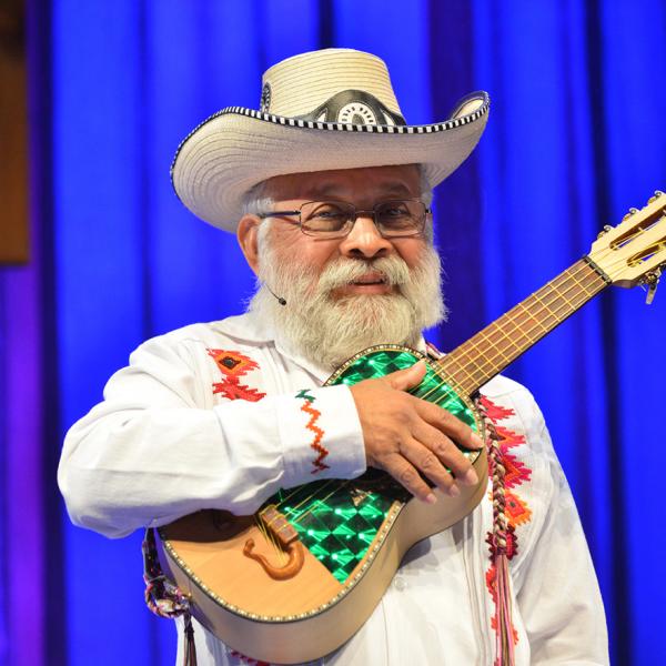 Musician with white beard in white suit and hat holding small guitar. 