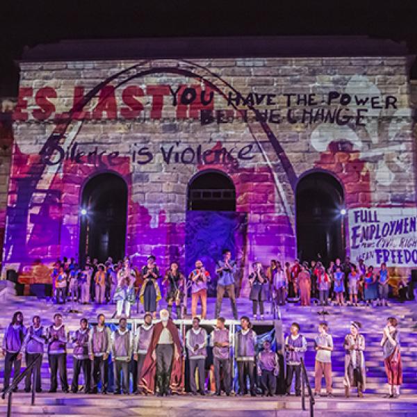 Performers line the steps of s stone building with art projections on it at night