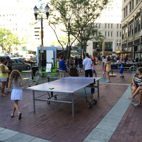 People on the sidewalk of a city doing hula hoop near a ping-pong table. 