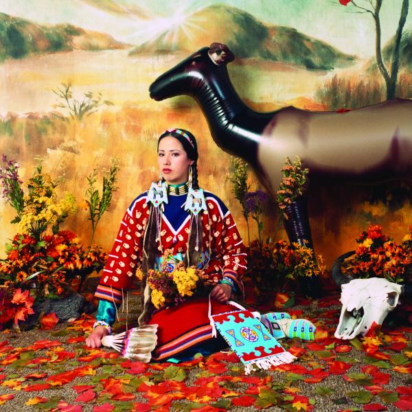 Woman in Native outfit sitting in front of a plains background  with a cow skull next to her and an inflatable elk behind her. 