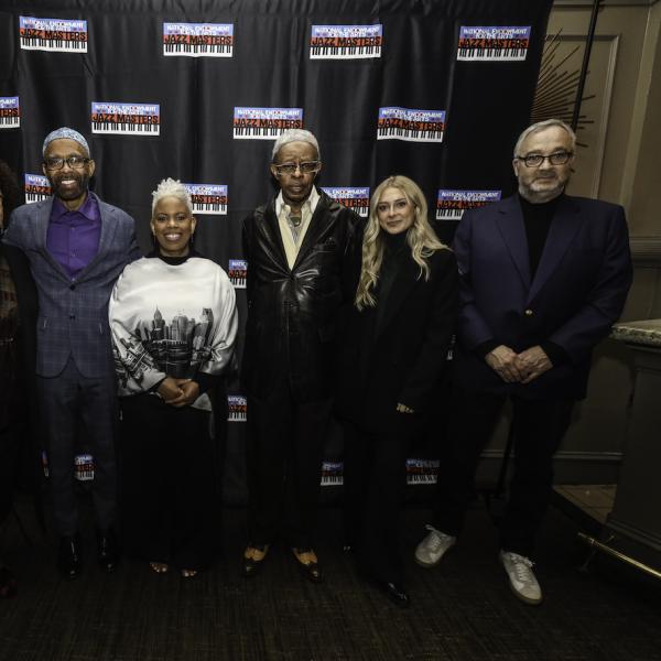 A group of six people stand smiling at the camera in front of a background with the National Endowment for the Arts Jazz Masters logo repeated on it. 