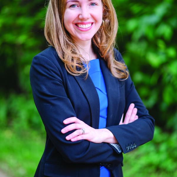 White woman wearing a blue dress and a dark blue blazer, standing in front of a wooded backround