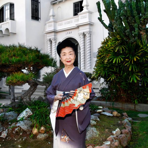 A Japanese woman holding a fan poses for a photo in front of a large white home