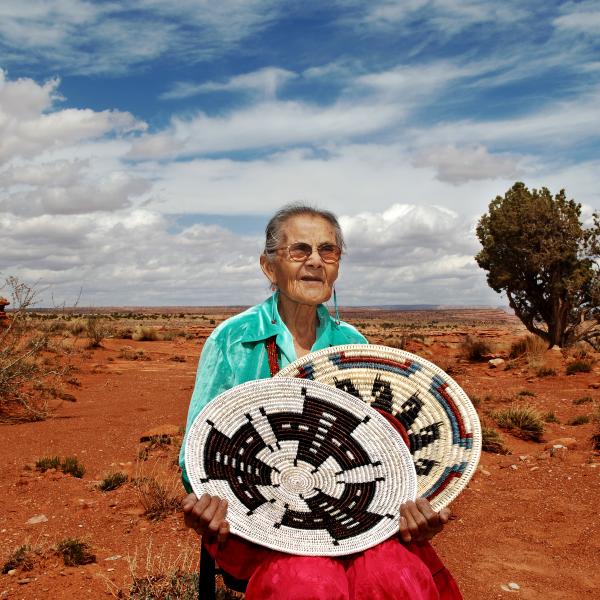 An elderly woman with glasses sits in the high desert holding two handwoven Navajo baskets. The red clay soil stands against the stark landscape and bright sky streaked with clouds. 