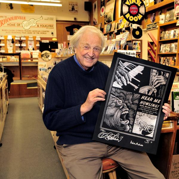 Man with white hair holding a poster sitting on a stool in a music shop with records and books. 