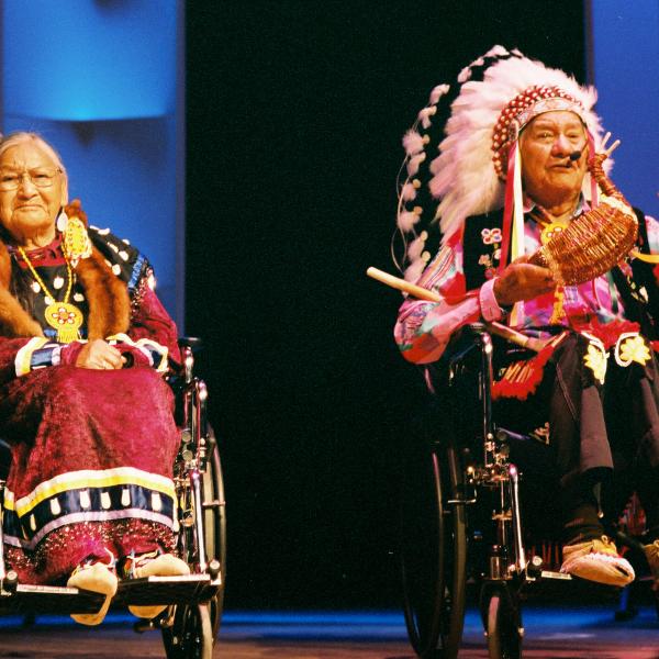 Older Native couple in wheelchairs on stage dressed in traditional Native clothing. 