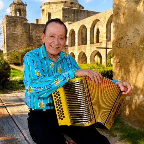 A man in a Southwestern blue printed shirt sits outside holding a gold-colored button accordion on his lap.  