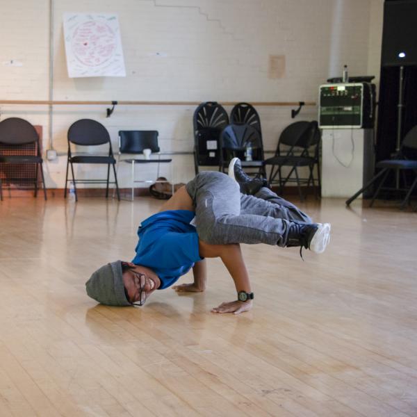 Student wearing hat dancing while a girl sitting cross-legged nearby watches. 