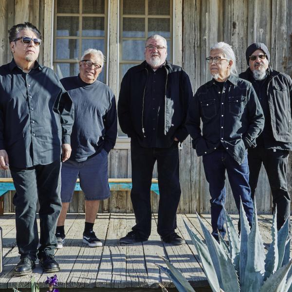 Five men wearing black stand on the porch of a weathered wooden building with a cactus in the foreground