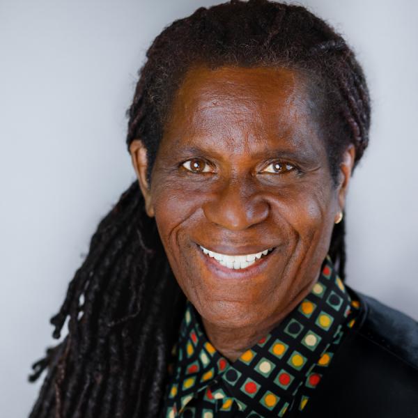A man with long dark hair wears a colorful shirt and black vest is smiling
