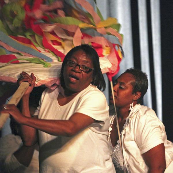 Three women wearing white clothes carry large colorful decorations