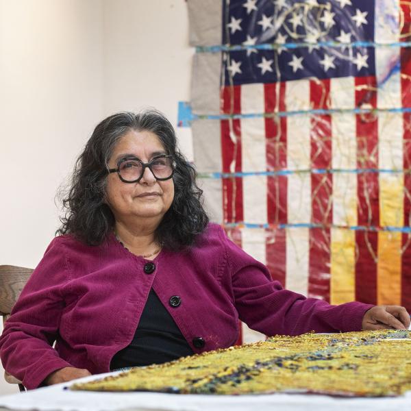 Hispanic/Latinx woman wearing black glasses and a purple cardigan sitting at a table with her artwork. An artwork featuring the American flag is behind her on the right. 