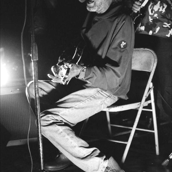 A black and white photo of an older man wearing a baseball-style hat, sitting on a folding chair playing the guitar and singing into a microphone. You can see another person in the background also playing music