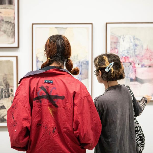 Two people with their backs to the viewer stand looking at a series of art works hung on a wall in a gallery