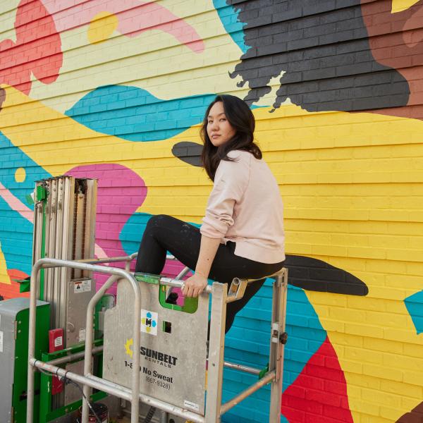 Asian women on scaffolding in front of mural she is painting. 