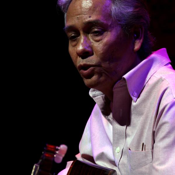 Man in white shirt with white hair brushed back playing guitar on stage. 