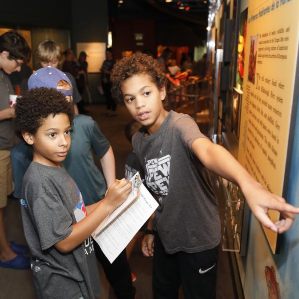 Two boys engage in an art exhibit at a museum. In the foreground, one child pointing at an information board while another take notes. 