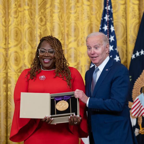 Older white male in blue suit posing with Black woman in red dress in front of flags and gold curtain.