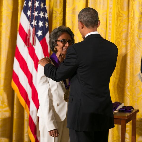 Woman in white suit wearing glasses accepting medal from Black man in dark suit in front of flags and gold curtain
