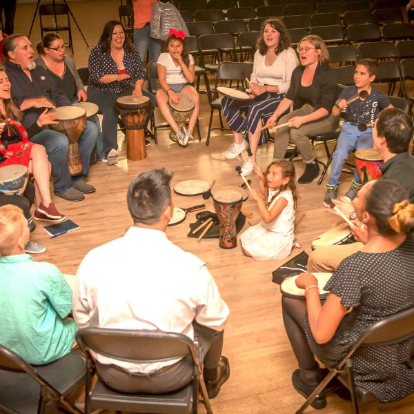 Large indoor drum circle of men and women with a small child in the middle on the floor m