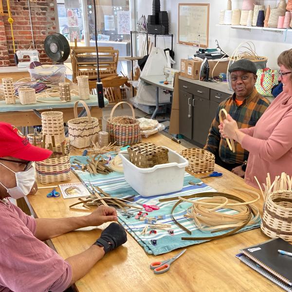 A small group of adults sit at a work table creating medium-sized baskets in an indoor studio setting with natural light.