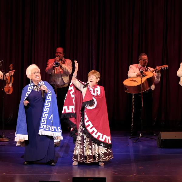 Two older women-one wearing a blue cape and one wearing a red cape-sing on stage with a mariachi band in the background