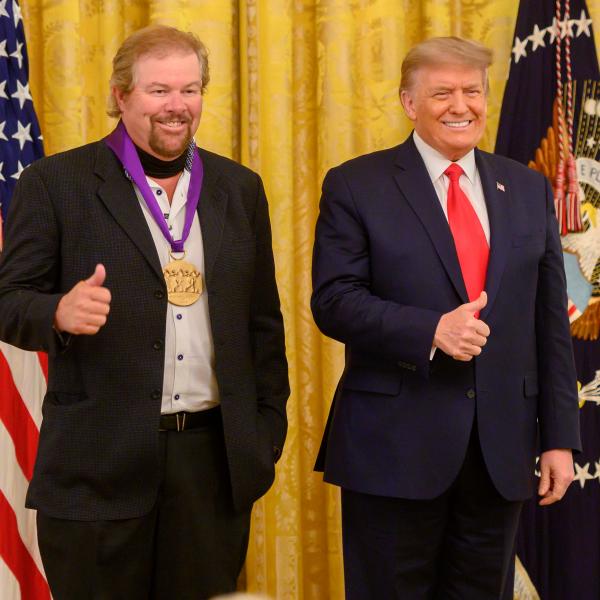 Two men standing in front of US flag holding thumbs up.