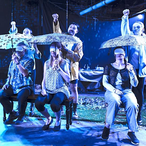 Four individuals sit in chairs holding umbrellas and wearing blindfolds. Behind them, four people stand with their hands raised, prepared to drop something onto the umbrella. 