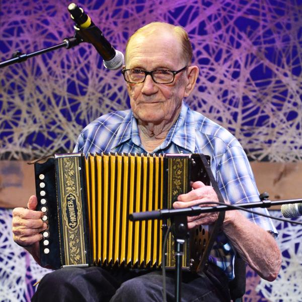 An elder man with glasses sits playing a button accordion with microphones around him and a colorful backdrop 
