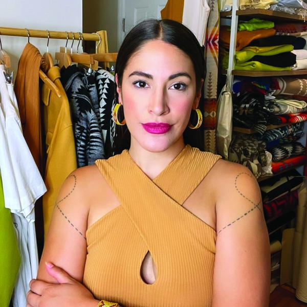 Woman with hair pulled back wearing a sleeveless yellow dress posing in front of a studio full of clothing. 