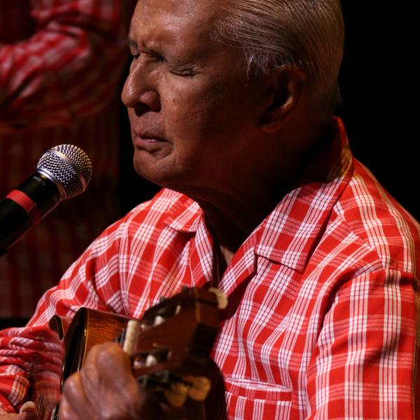 Balding Hawaiian man in red checkered shirt playing a uke. 