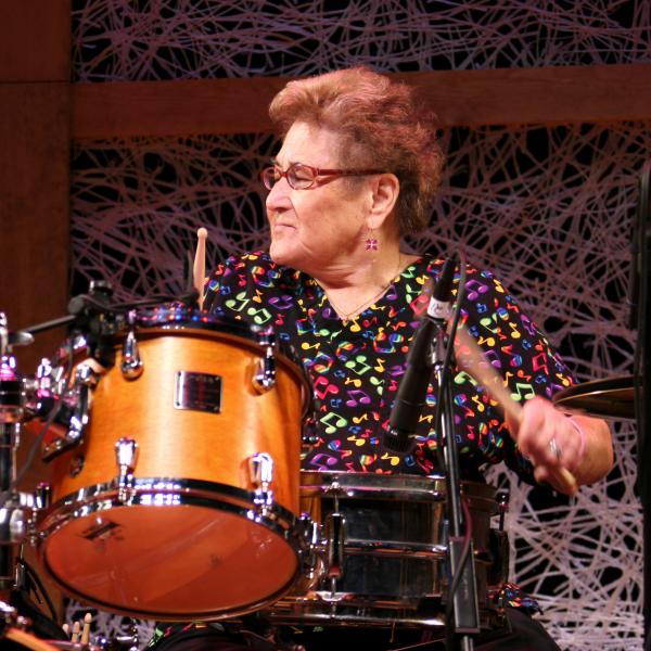 Older woman with short brown hair in colorful shirt playing drums. 