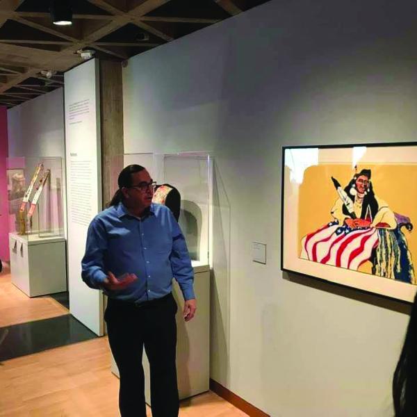 Man in blue shirt standing in front of painting in museum and gesturing while talking. 