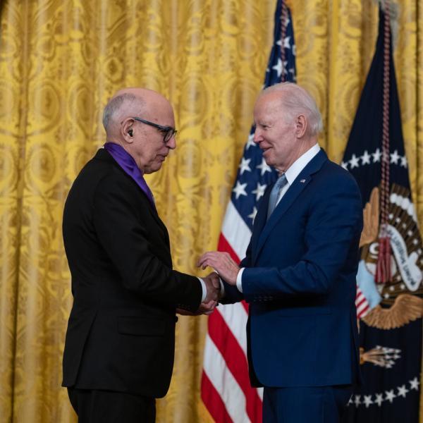 Older white male in blue suit posing with white man wearing glasses in black suit in front of flags and gold curtain.