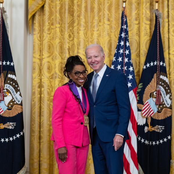 Older white male in blue suit posing with Black woman in pink outfit in front of flags and gold curtain.