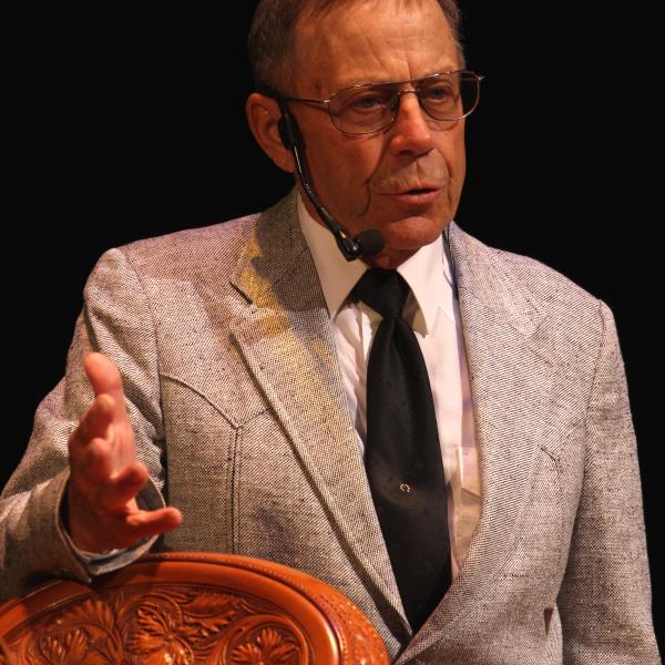 White man in light suit and dark tie wearing glasses with short hair, standing next to a saddle. 