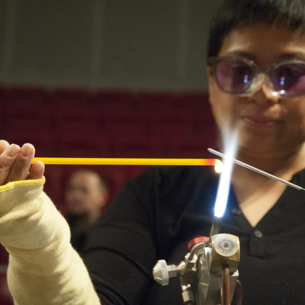 Person wearing goggles holds a yellow glass rod over open flame