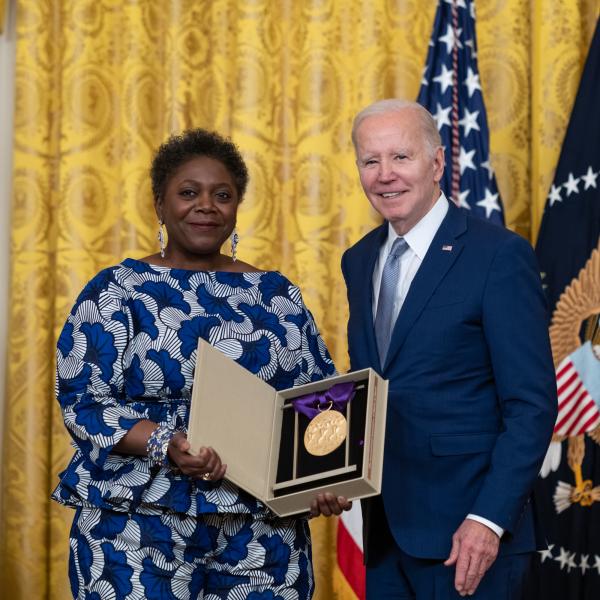 Older white male in blue suit posing with Black woman in blue floral dress in front of flags and gold curtain. 