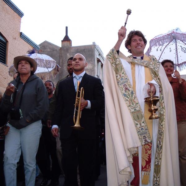 Man with trumpet next to priest blessing crowd in parade down street.